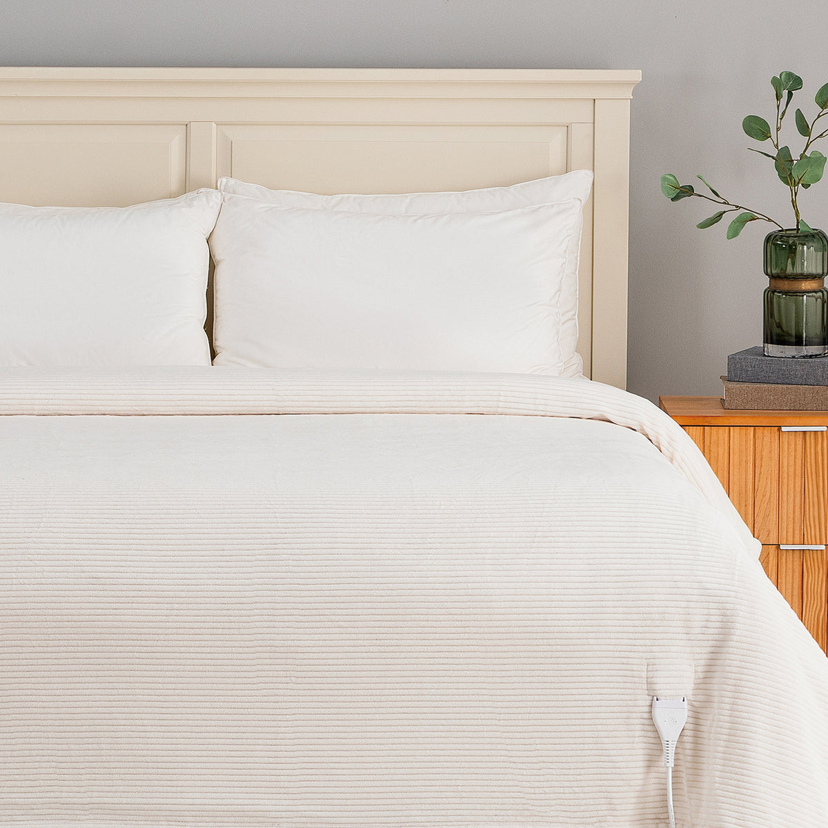A bed dressed in white ribbed bedding, including a duvet cover and two pillows, set against a light wood headboard. A wooden nightstand with a green glass vase is visible on the right against a neutral gray wall.