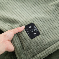 Close-up of a ribbed knit garment in muted sage green with a black built-in control patch held by a hand. The patch shows a digital display and icons for power, temperature, and heating modes.