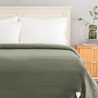 An olive green ribbed blanket or duvet cover spread across a bed with white pillows and a light wood headboard, shown in a styled bedroom setting next to a wooden nightstand with a green glass vase.