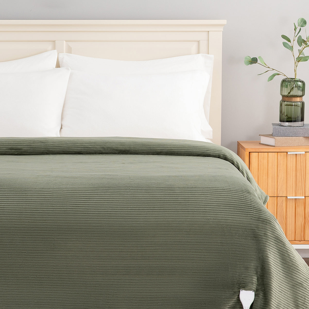 An olive green ribbed blanket or duvet cover spread across a bed with white pillows and a light wood headboard, shown in a styled bedroom setting next to a wooden nightstand with a green glass vase.