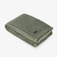 An olive green corduroy throw blanket, folded neatly, featuring a small black fabric brand tag on the side, set against a clean white background.