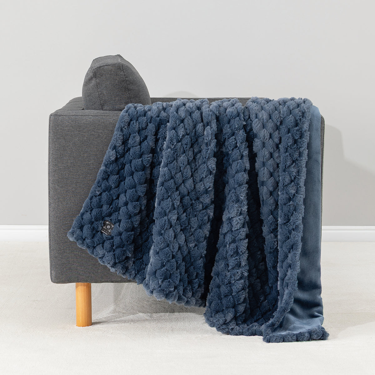 A textured navy blue throw blanket with a chunky knit pattern and a smooth velvet reverse, draped over a modern gray armchair with a light wood leg against a neutral studio background.