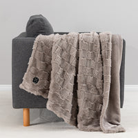 A plush taupe throw blanket with a textured basketweave pattern and fringed edges draped over a gray armchair, with a matching solid taupe backing visible on the right side.