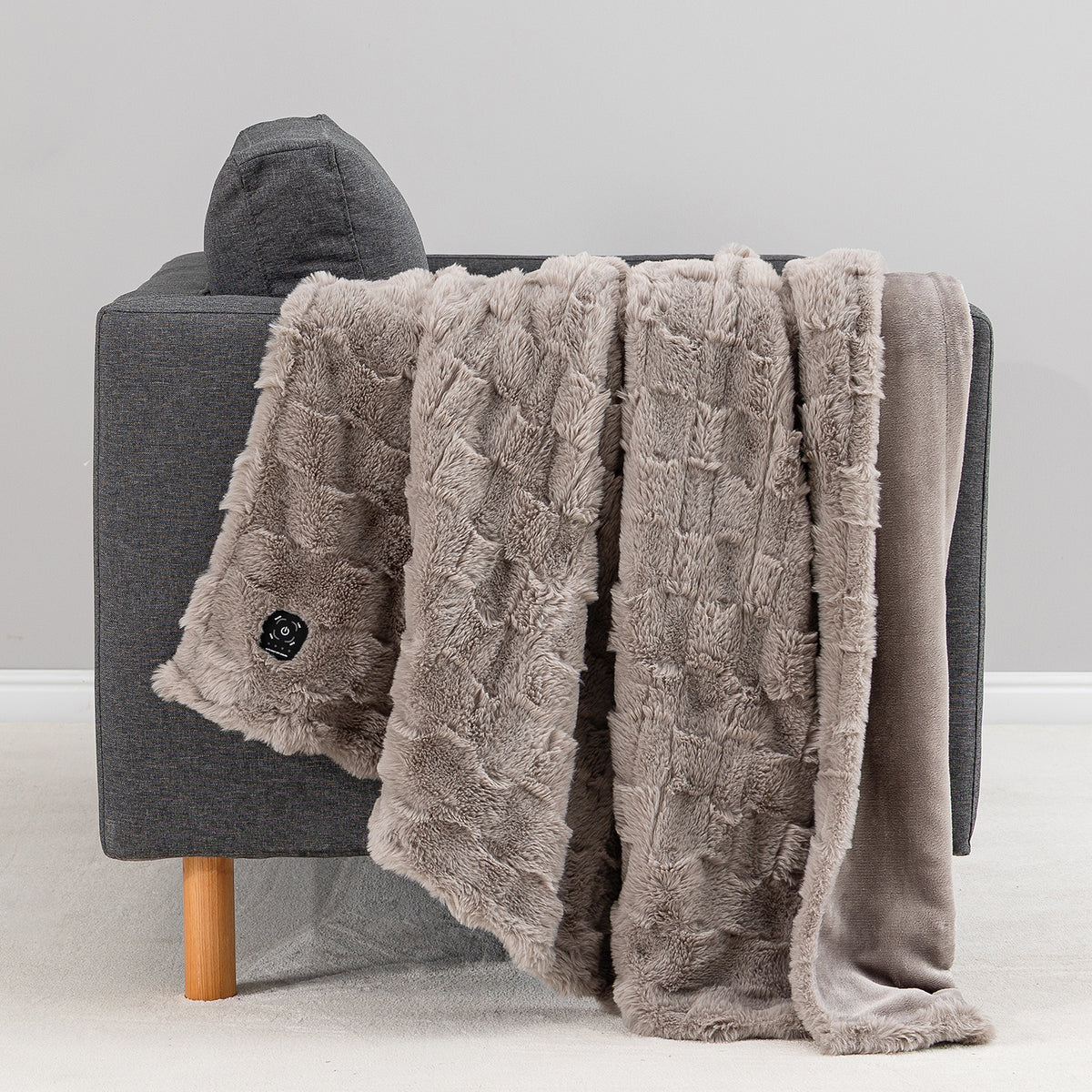 A plush taupe throw blanket with a textured basketweave pattern and fringed edges draped over a gray armchair, with a matching solid taupe backing visible on the right side.