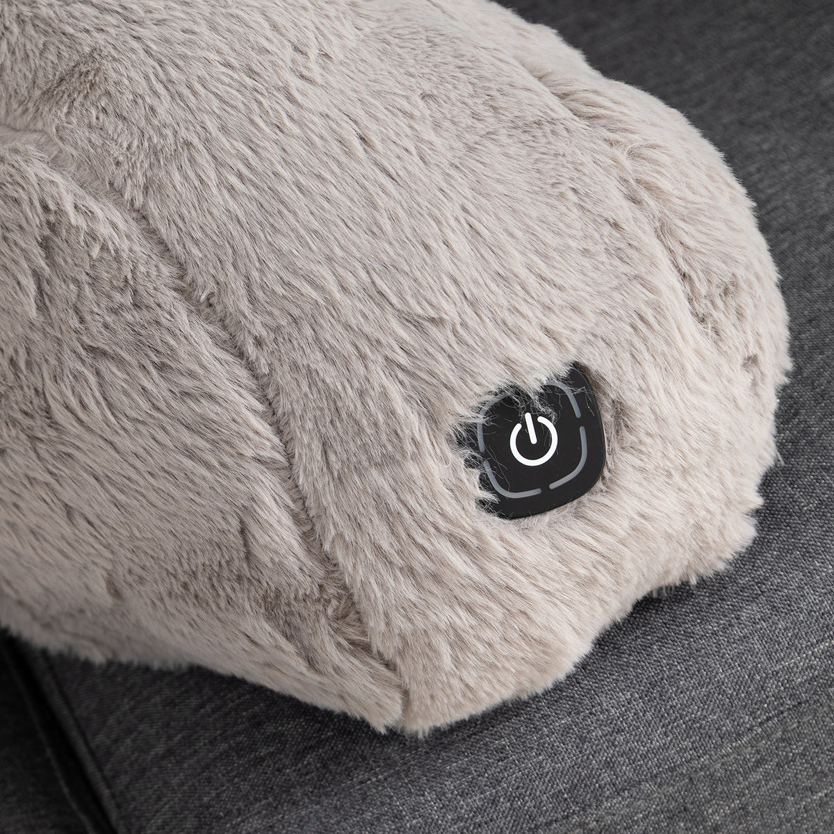 Close-up of a fluffy light gray heated massage pillow with a black power button on a dark gray fabric surface.
