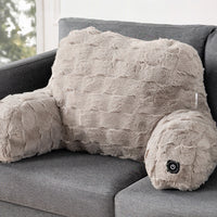 A plush, furry massage pillow shaped like a human body with arm and headrest, resting on a gray fabric sofa. The pillow is a light taupe-beige color with a small black power button on the headrest.