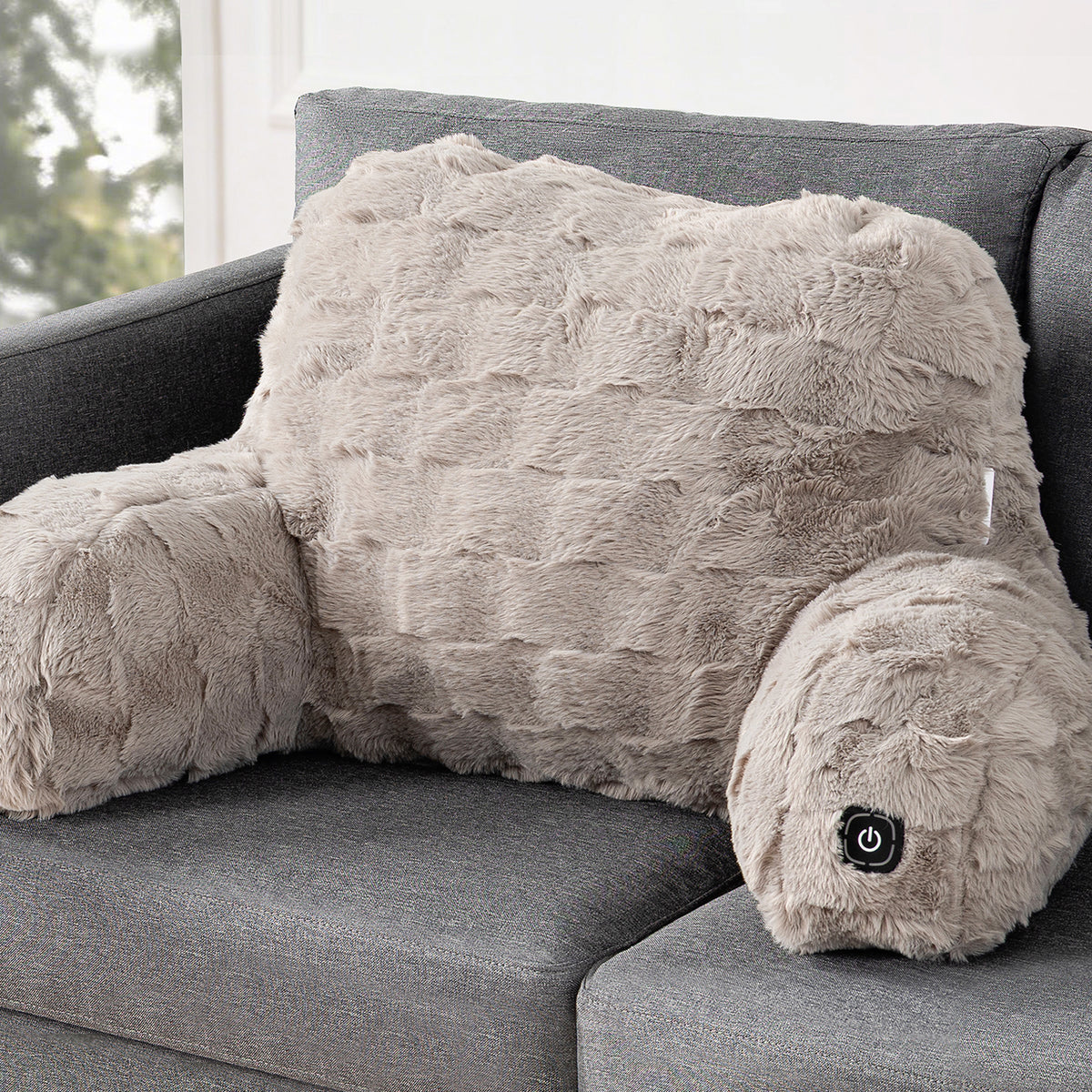 A plush, furry massage pillow shaped like a human body with arm and headrest, resting on a gray fabric sofa. The pillow is a light taupe-beige color with a small black power button on the headrest.