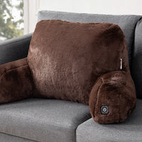 A brown plush heated pillow with armrests, resting on a gray fabric sofa. The pillow features a power button on the lower right armrest.