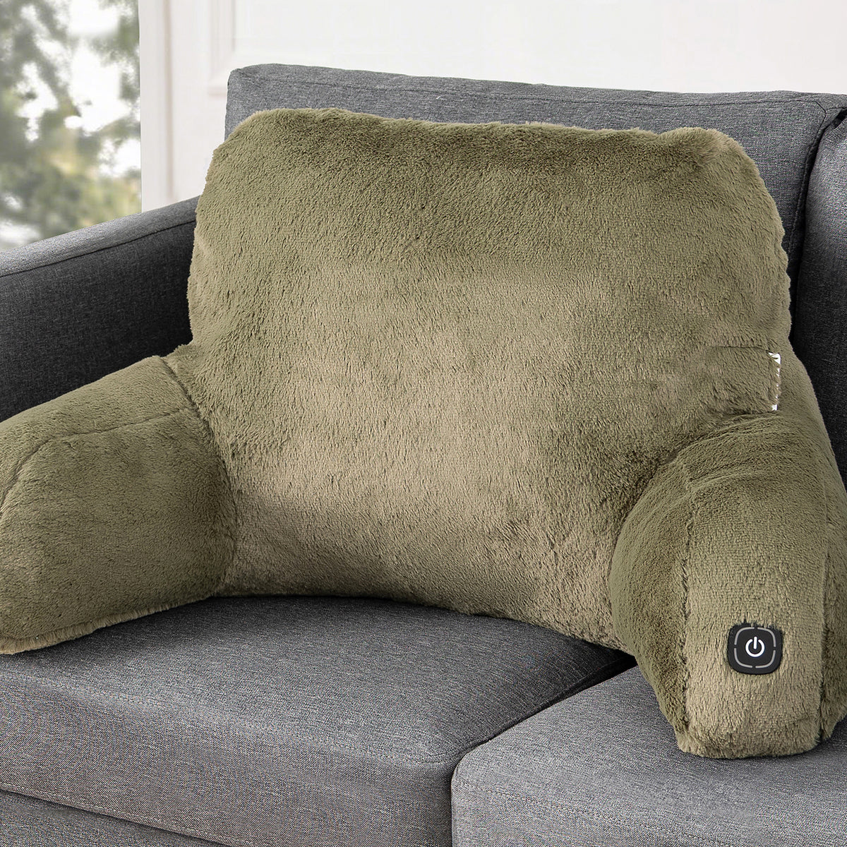 An olive green plush heated corner support pillow resting on a gray fabric sofa, with a small power button visible on the lower right armrest.