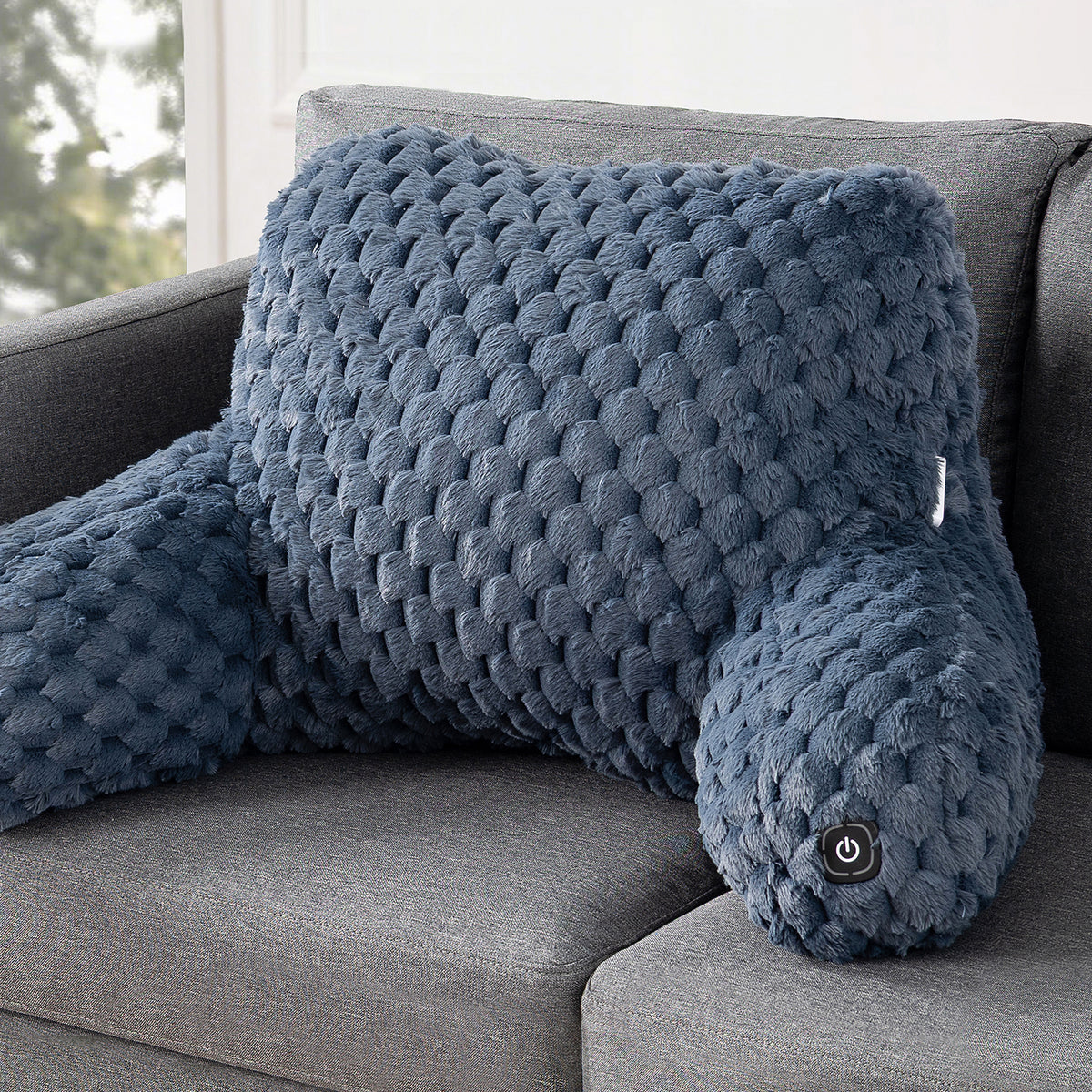 A textured, contoured lumbar pillow in a slate blue color, resting on a gray fabric sofa. The pillow features a raised, bumpy circular pattern and a small power button on the head.