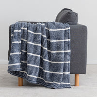 A textured throw blanket with horizontal navy and white stripes draped over a dark gray armchair with light wood legs, set against a neutral gray background.