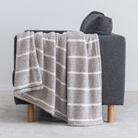 A plush throw blanket with horizontal gray and white stripes draped over a dark gray armchair with light wood legs, set against a neutral light gray background.