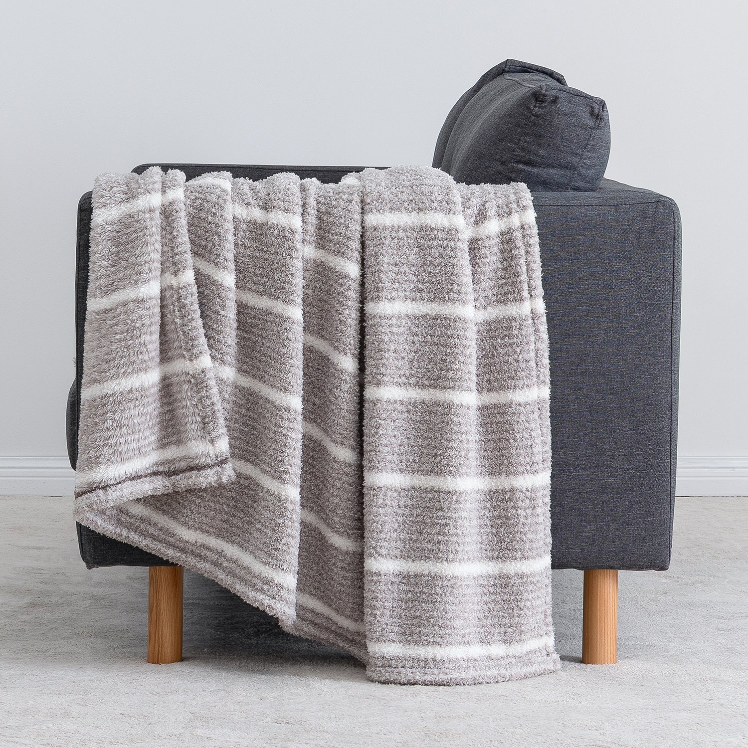 A plush throw blanket with horizontal gray and white stripes draped over a dark gray armchair with light wood legs, set against a neutral light gray background.