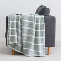 A textured throw blanket with horizontal white stripes in a muted sage green, draped over the arm of a dark gray armchair with light wood legs.