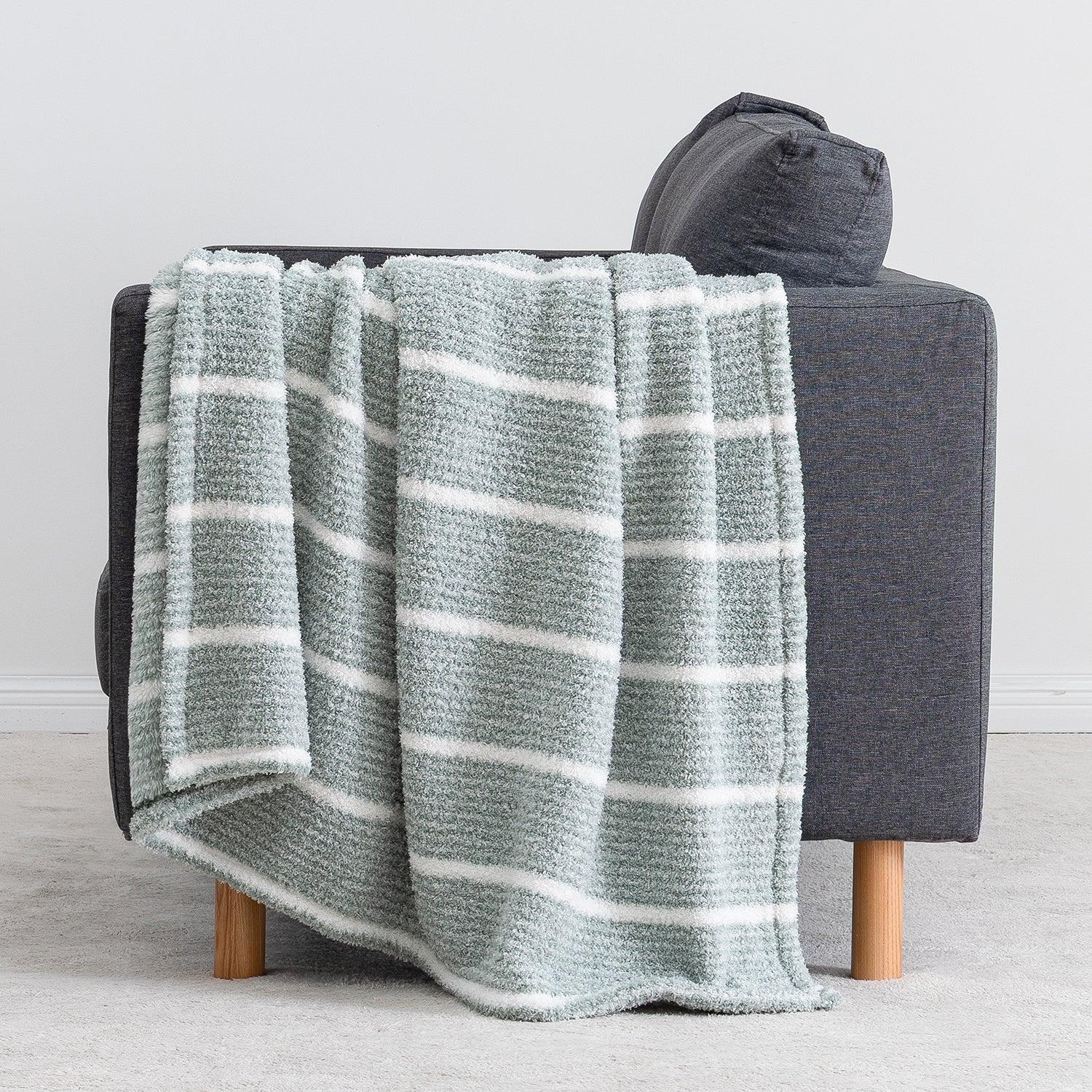 A textured throw blanket with horizontal white stripes in a muted sage green, draped over the arm of a dark gray armchair with light wood legs.