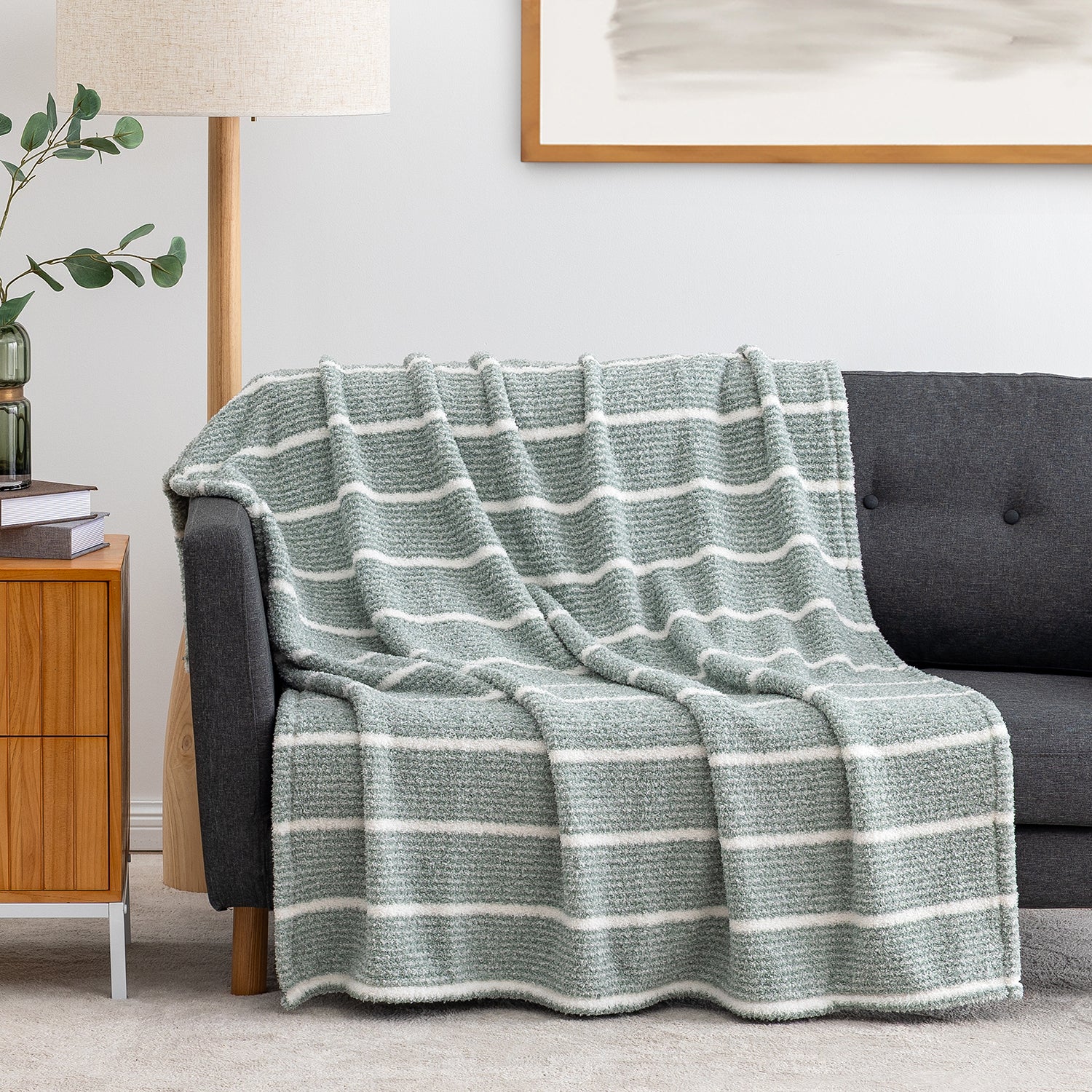 A textured knit throw blanket with horizontal white stripes in a muted sage green, draped over a dark gray sofa in a modern living room setting.