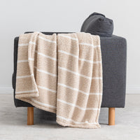 A beige throw blanket with white stripes draped over a dark gray armchair with light wood legs, set against a neutral gray wall and floor.