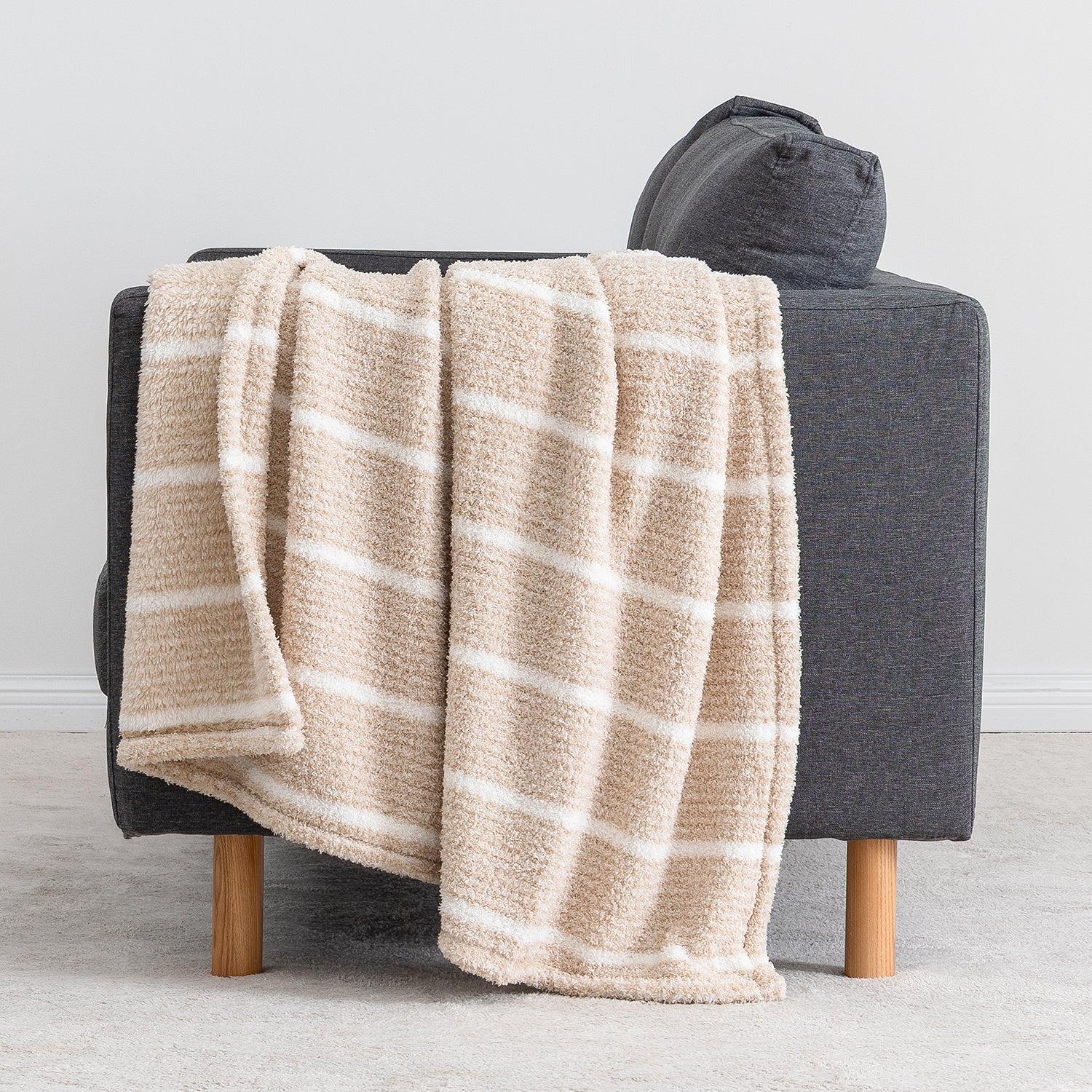 A beige throw blanket with white stripes draped over a dark gray armchair with light wood legs, set against a neutral gray wall and floor.