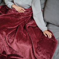 A woman sitting on a grey sofa with a red velvetloft throw blanket draped over her legs.