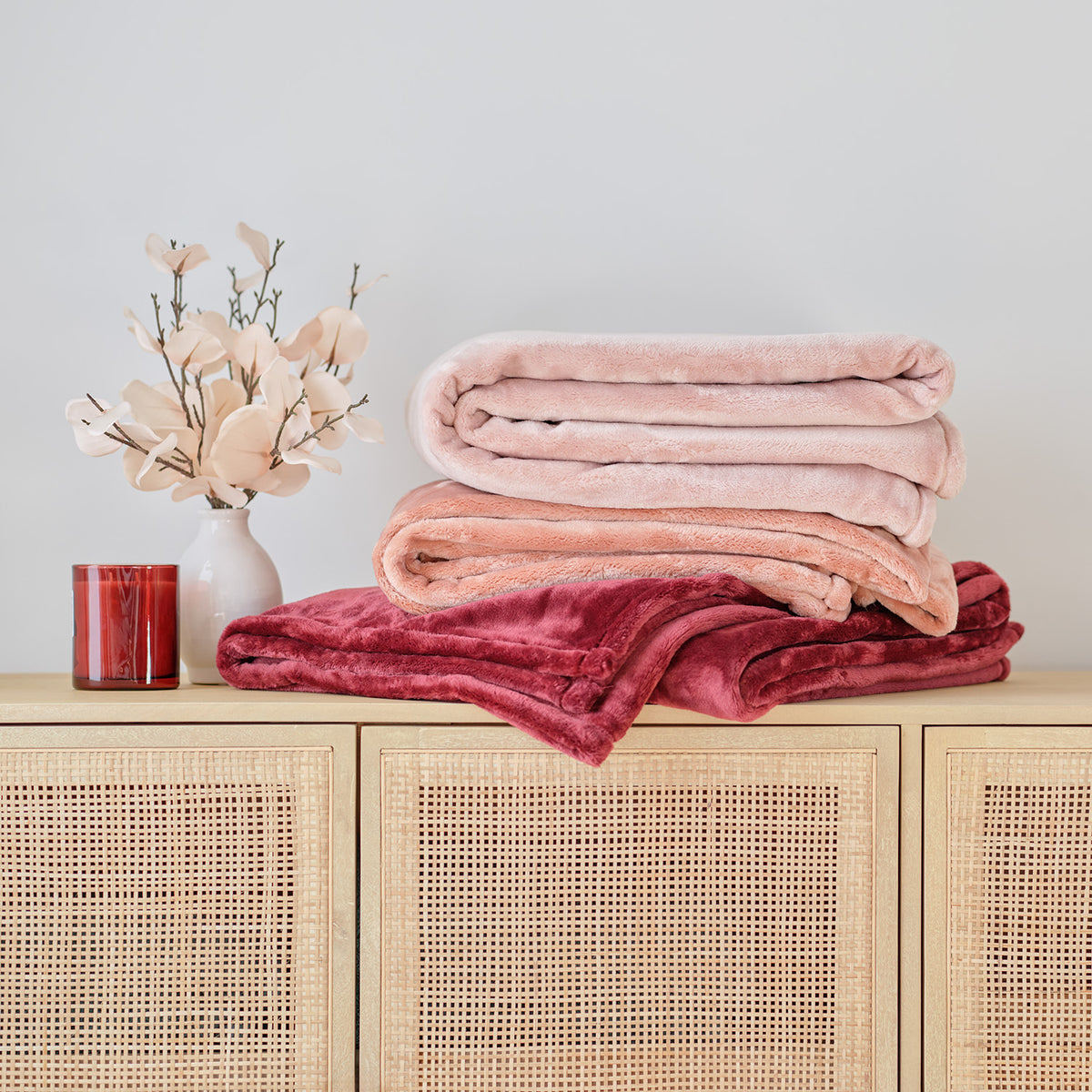 A stack of 3 rose colored Premium VelvetLoft Throws stacked on a boho console table with a bouquet and candle.