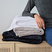 A woman sitting next to a stack of 3 neutral colored throw blankets while resting her arm on them.