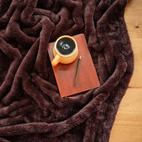A brown Premium Extra-Fluffy Throw laid on a wooden floor with a coffee, notebook, and pen.