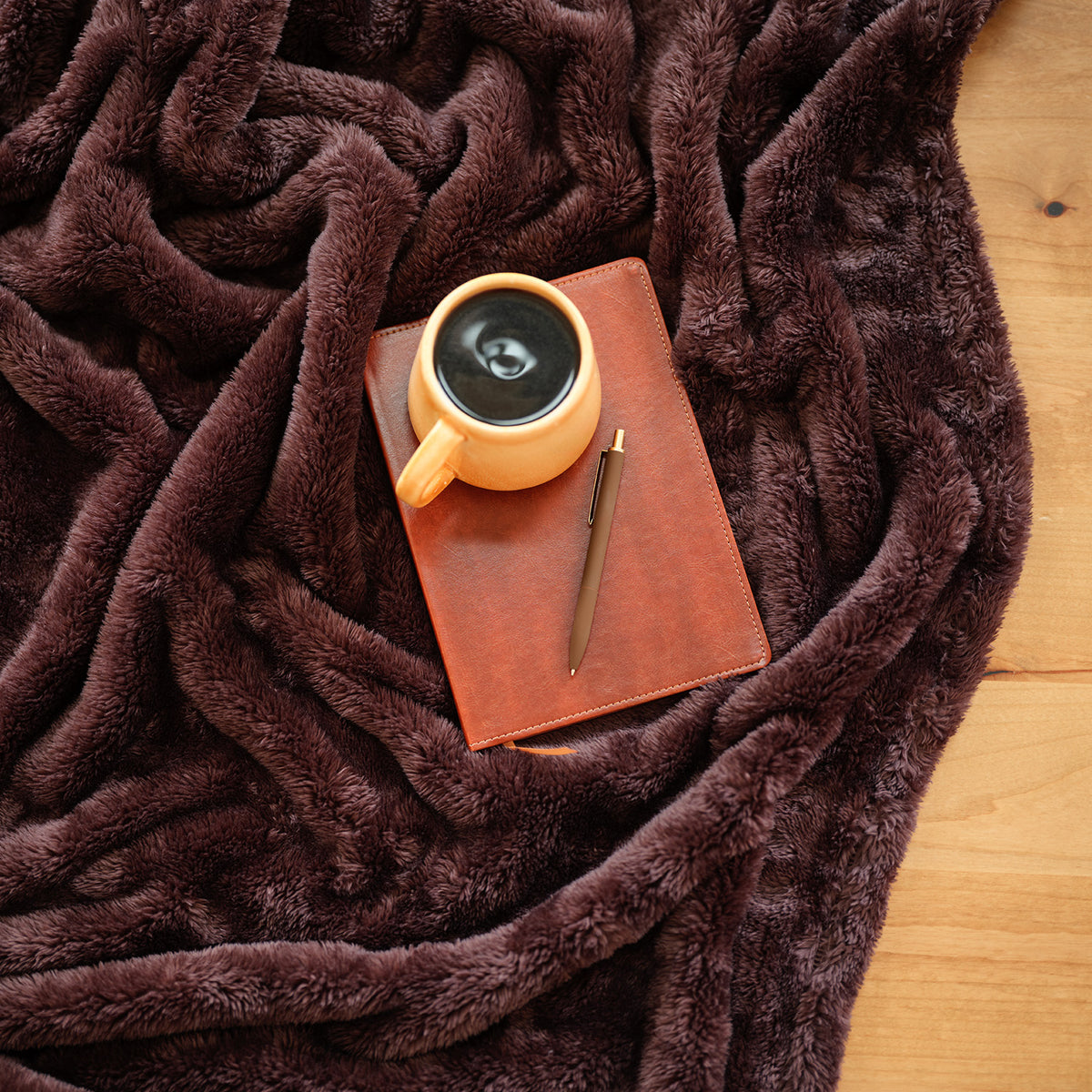 A brown Premium Extra-Fluffy Throw laid on a wooden floor with a coffee, notebook, and pen.