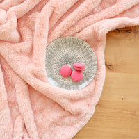 A pink Premium Extra-Fluffy Throw laid on a wooden floor next to a plate of pink macaroons.