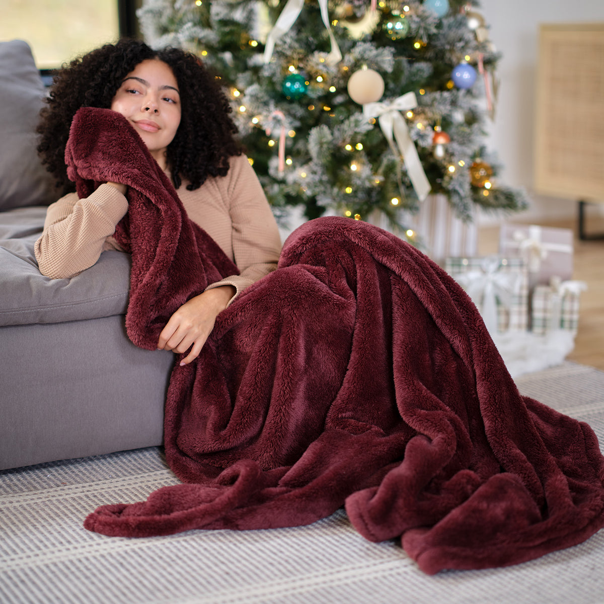 A woman sitting on the floor next to a christmas tree with a maroon Premium Extra-Fluffy Throw.