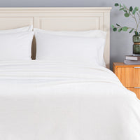 White bedding set including a flat sheet and two pillowcases on a bed with a cream-colored wooden headboard, next to a wooden nightstand with a green vase.