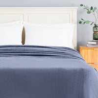 A navy blue textured blanket is neatly spread across a bed with white pillows and a light wood headboard, set in a bedroom next to a wooden nightstand with a green plant.