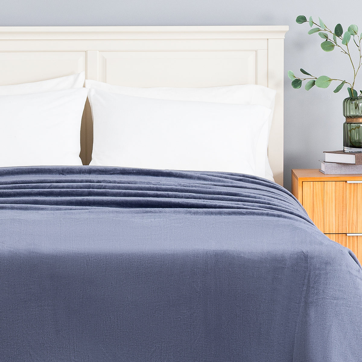 A navy blue textured blanket is neatly spread across a bed with white pillows and a light wood headboard, set in a bedroom next to a wooden nightstand with a green plant.