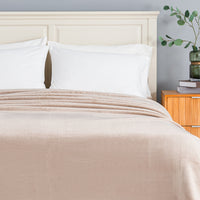 A beige textured blanket is neatly spread across a bed with white pillows and a cream-colored wooden headboard, set against a gray wall with a wooden nightstand on the right.