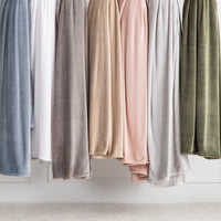 A row of plush velvet throw blankets in muted neutral and pastel colors hanging side by side against a white wall.