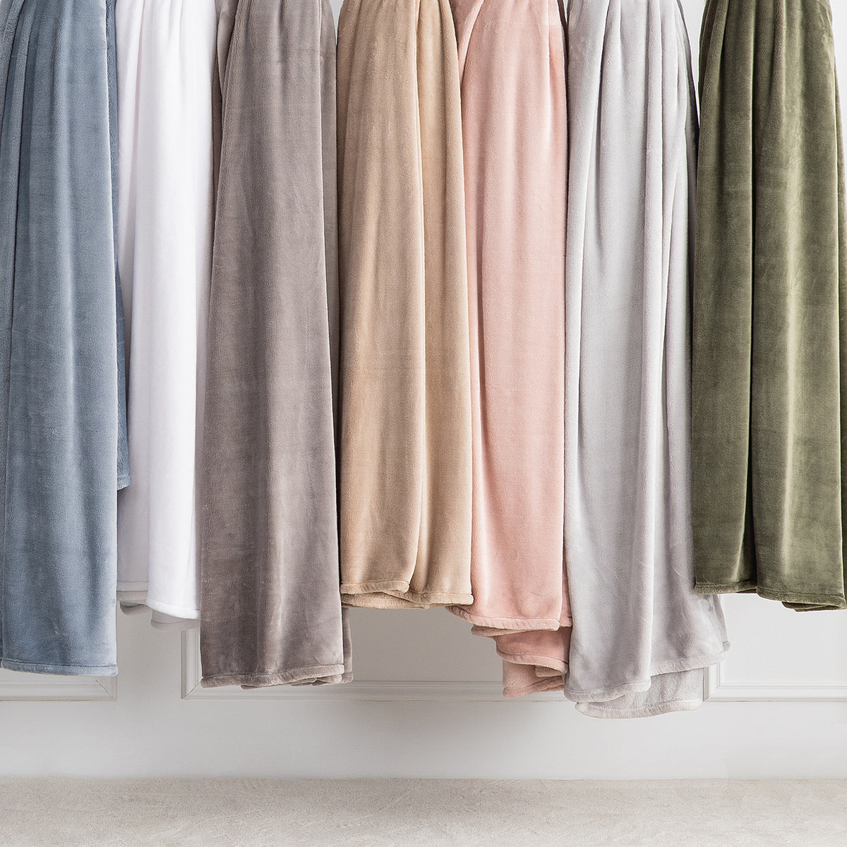 A row of plush velvet throw blankets in muted neutral and pastel colors hanging side by side against a white wall.