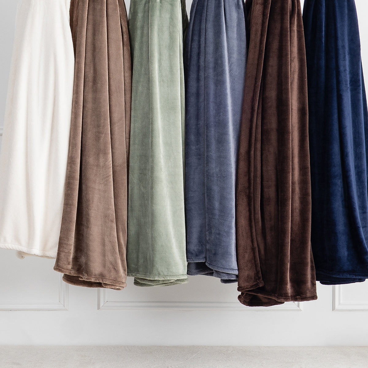 Five plush velvet throw blankets in assorted colors including cream, taupe, sage green, slate blue, and navy blue, hanging side-by-side against a white paneled wall.