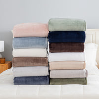 Two stacks of folded plush throw blankets in various colors are displayed on a white bed. The left stack features dusty pink, light blue, cream, taupe, light gray, and navy blue blankets.