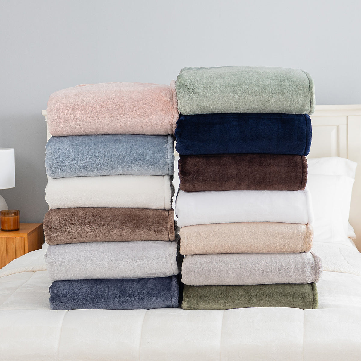 Two stacks of folded plush throw blankets in various colors are displayed on a white bed. The left stack features dusty pink, light blue, cream, taupe, light gray, and navy blue blankets.