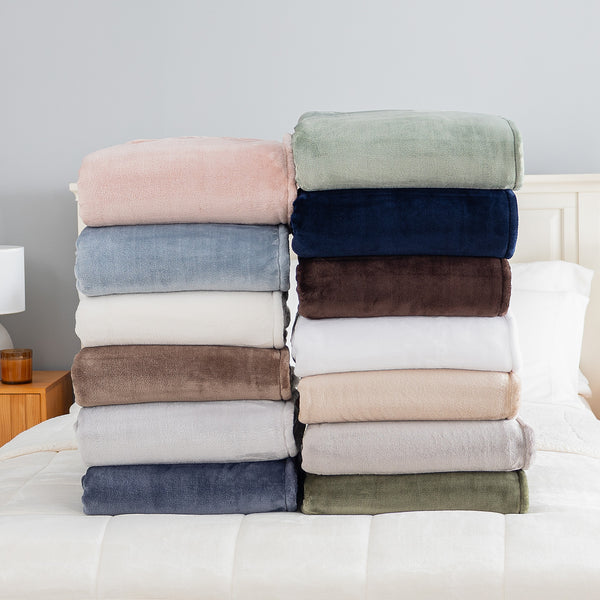 Two stacks of folded plush throw blankets in various colors are displayed on a white bed. The left stack features dusty pink, light blue, cream, taupe, light gray, and navy blue blankets.