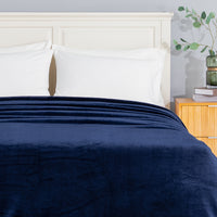 A navy blue plush blanket is neatly folded and spread across the foot of a bed with white pillows and a white headboard, next to a light wood nightstand with a green glass vase.