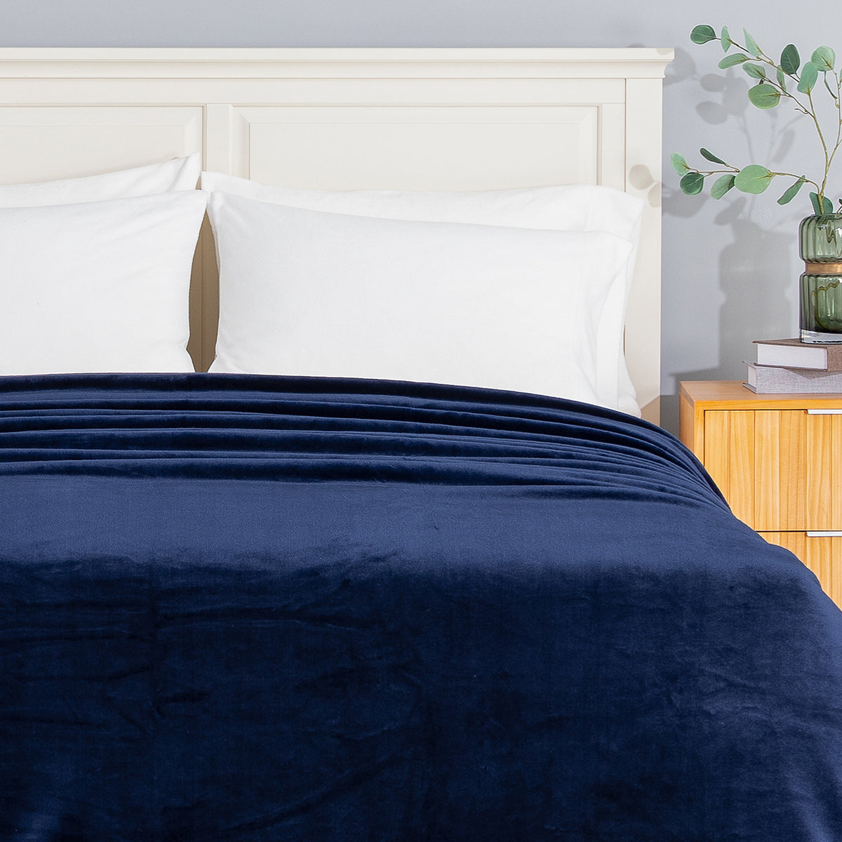 A navy blue plush blanket is neatly folded and spread across the foot of a bed with white pillows and a white headboard, next to a light wood nightstand with a green glass vase.