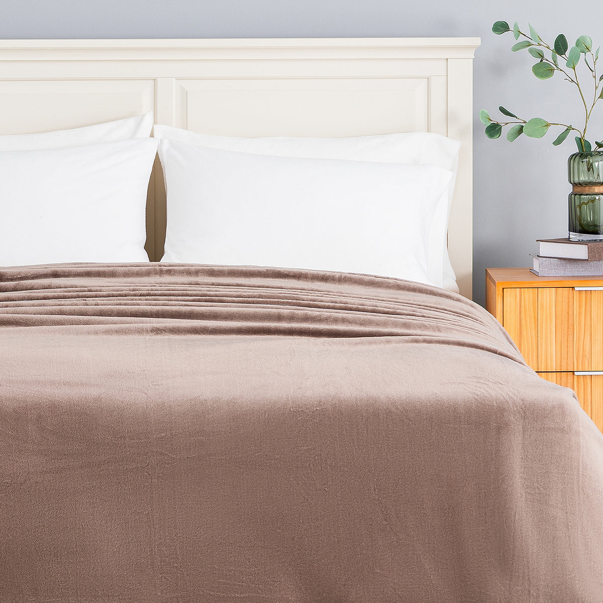A plush, soft brown fleece blanket is neatly spread across a bed with white pillows and a light wood headboard, set in a bedroom next to a wooden nightstand with a green plant.