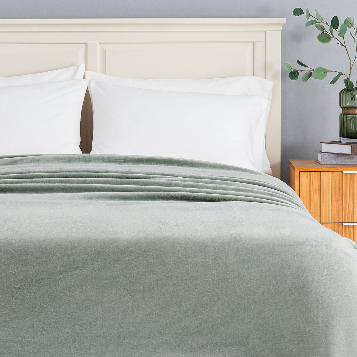 A sage green blanket is neatly spread across a bed with white pillows and a cream-colored wooden headboard, set in a bedroom next to a wooden nightstand with a green plant.