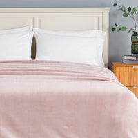 A dusty pink velvet throw blanket draped over a bed with white pillows and a cream-colored wooden headboard, set in a bedroom next to a wooden nightstand.