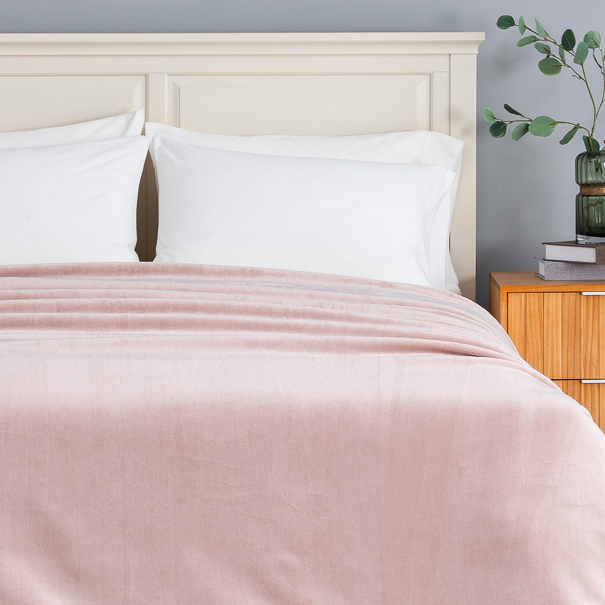 A dusty pink velvet throw blanket draped over a bed with white pillows and a cream-colored wooden headboard, set in a bedroom next to a wooden nightstand.