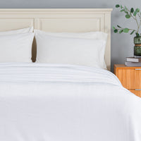 White bedding set with a duvet cover and two pillows neatly arranged on a bed with a cream-colored wooden headboard, next to a wooden nightstand with a green plant.