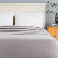 A bed dressed in white pillows and a light gray textured blanket, set against a cream-colored wooden headboard in a bedroom scene with a wooden nightstand and greenery.