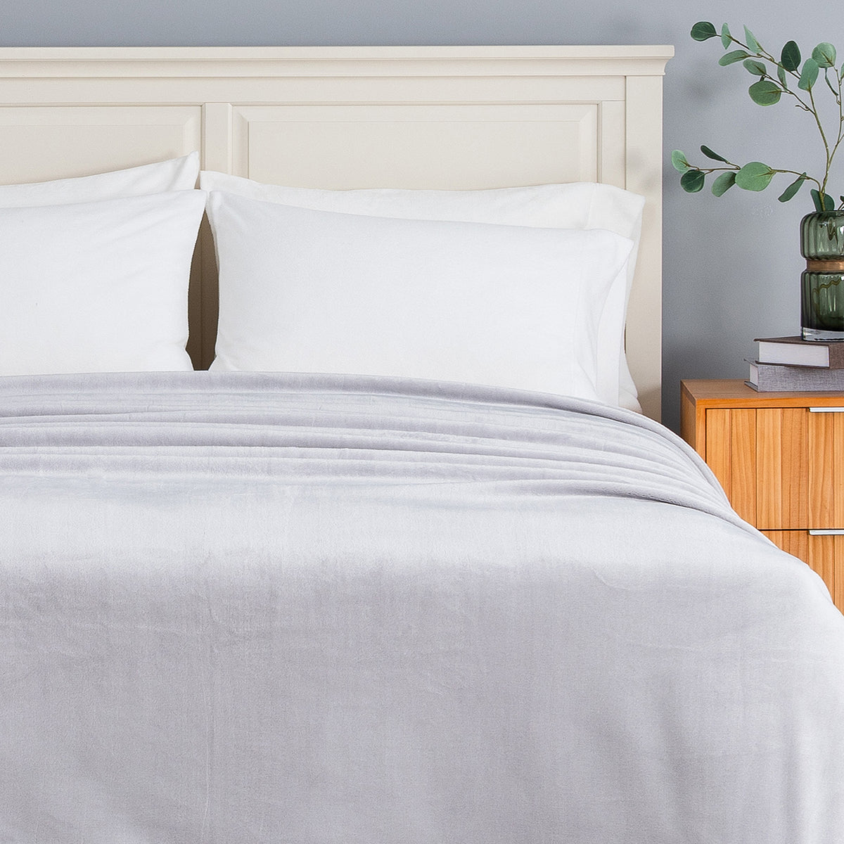 A light gray textured blanket and matching white pillowcases on a bed with a cream-colored wooden headboard, set against a gray wall with a wooden nightstand and greenery.