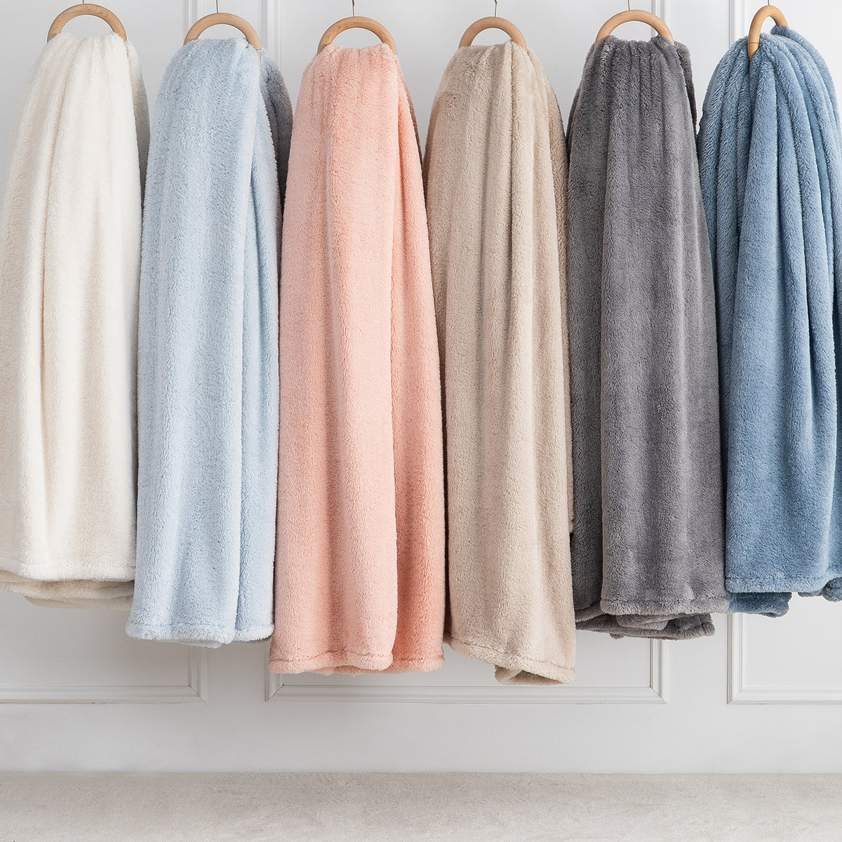 Premium Extra-Fluffy™ Blankets in six lighter Colors hanging side by side on a rail against a white paneled wall