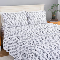 A bed dressed in a white bedding set with an all-over black Snoopy character print, shown in a styled bedroom with a light wood headboard, bedside table, books, and a plant.